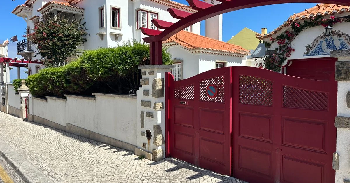 Charmante maison portugaise avec portail rouge et haies luxuriantes sous un ciel d'été limpide.
