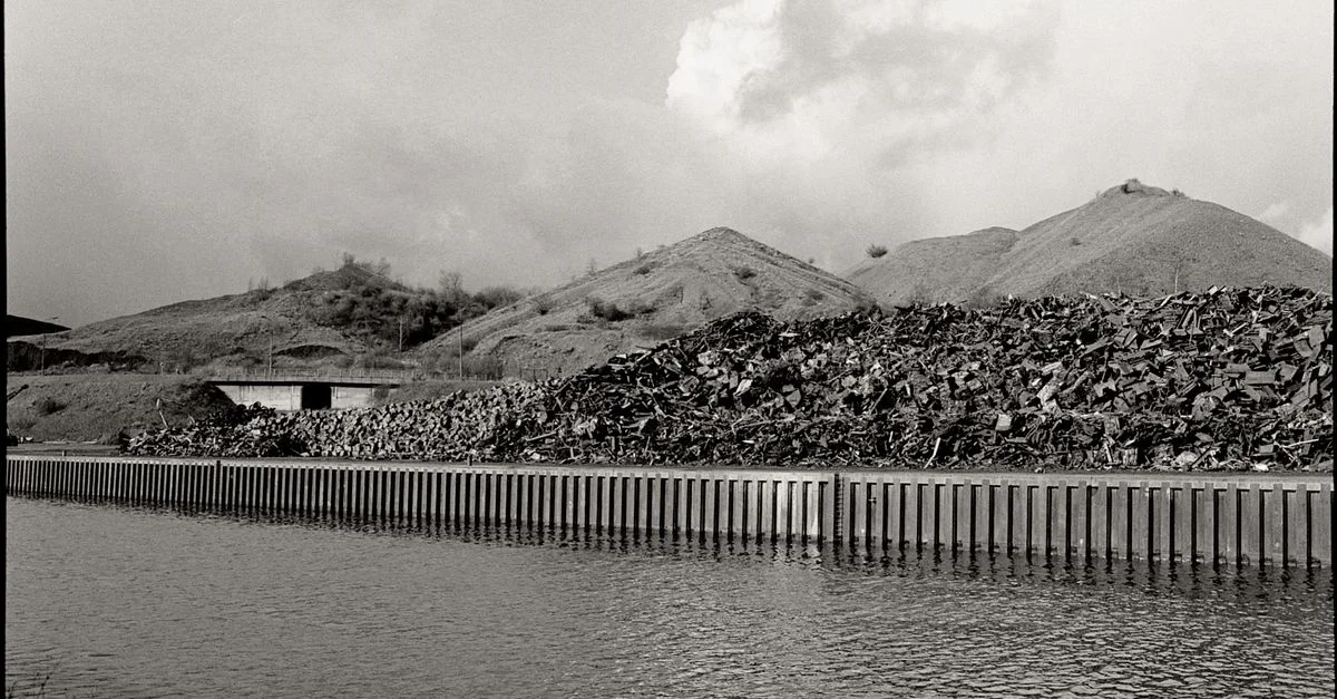 Image en niveaux de gris d'un canal industriel avec des tas de ferraille et des collines.